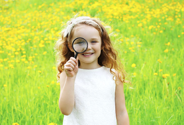 Smiling girl standing in a green field looking through a magnifying glass